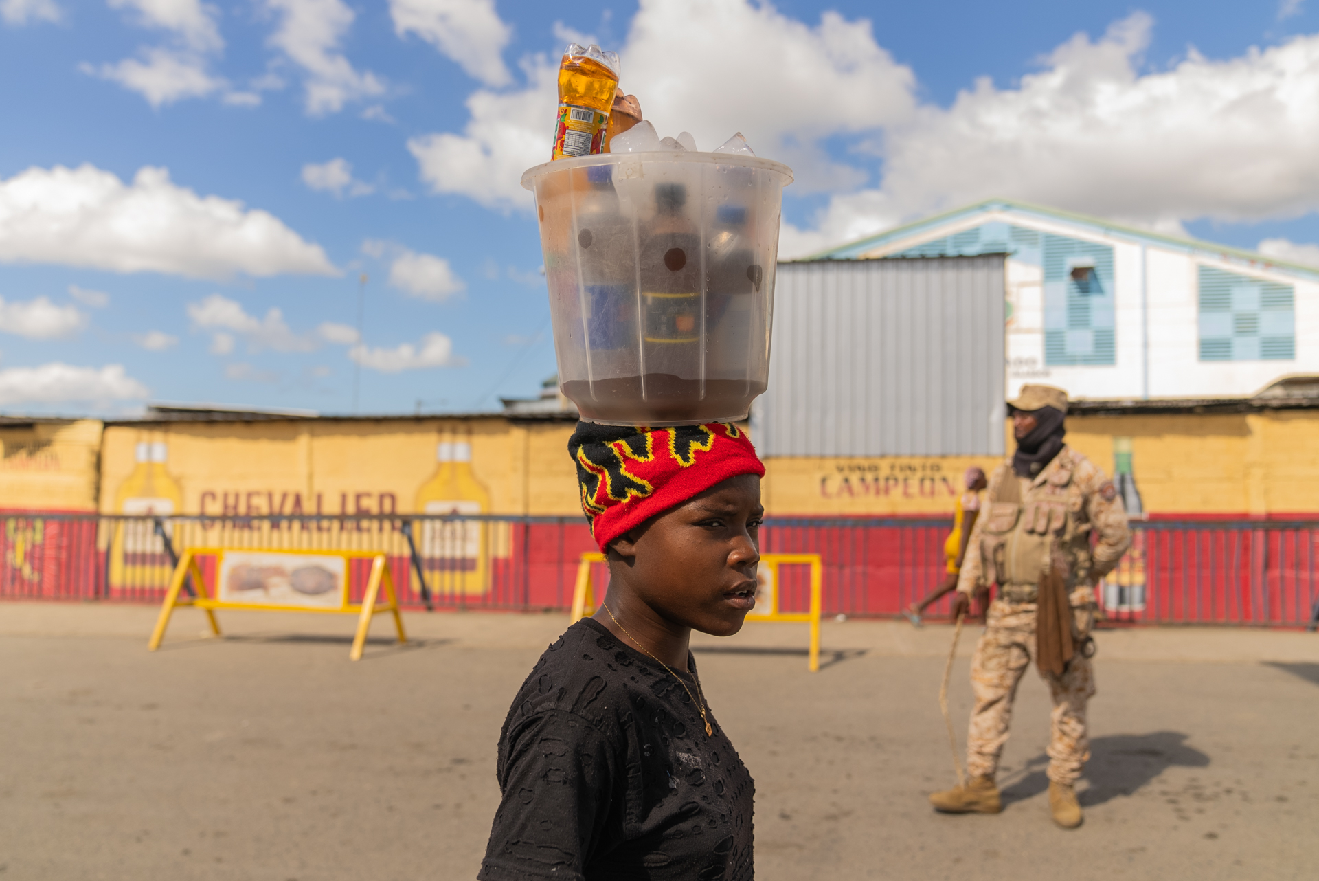 A young woman balances a large plastic bin filled with bottled beverages on her head at a border crossing, her expression focused and composed. An armed soldier in camouflage stands in the background; a painted wall reads "Chevalier.", a liquor advertisement