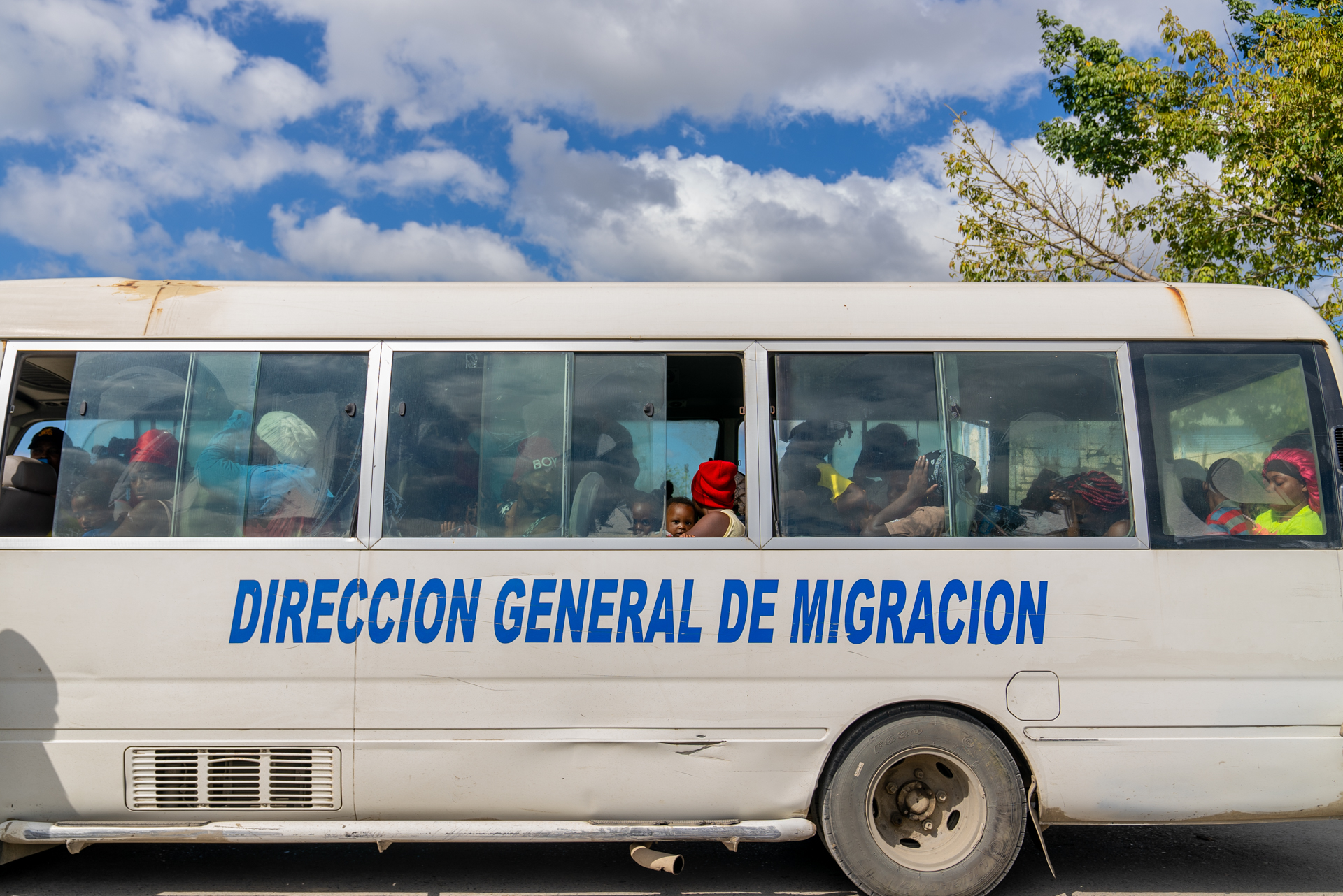 A crowded white bus marked Direccion General de Migracion is seen from the side, its windows filled with passengers including a small child peering out. The bus transports deportees to the Dominican border. data-caption=