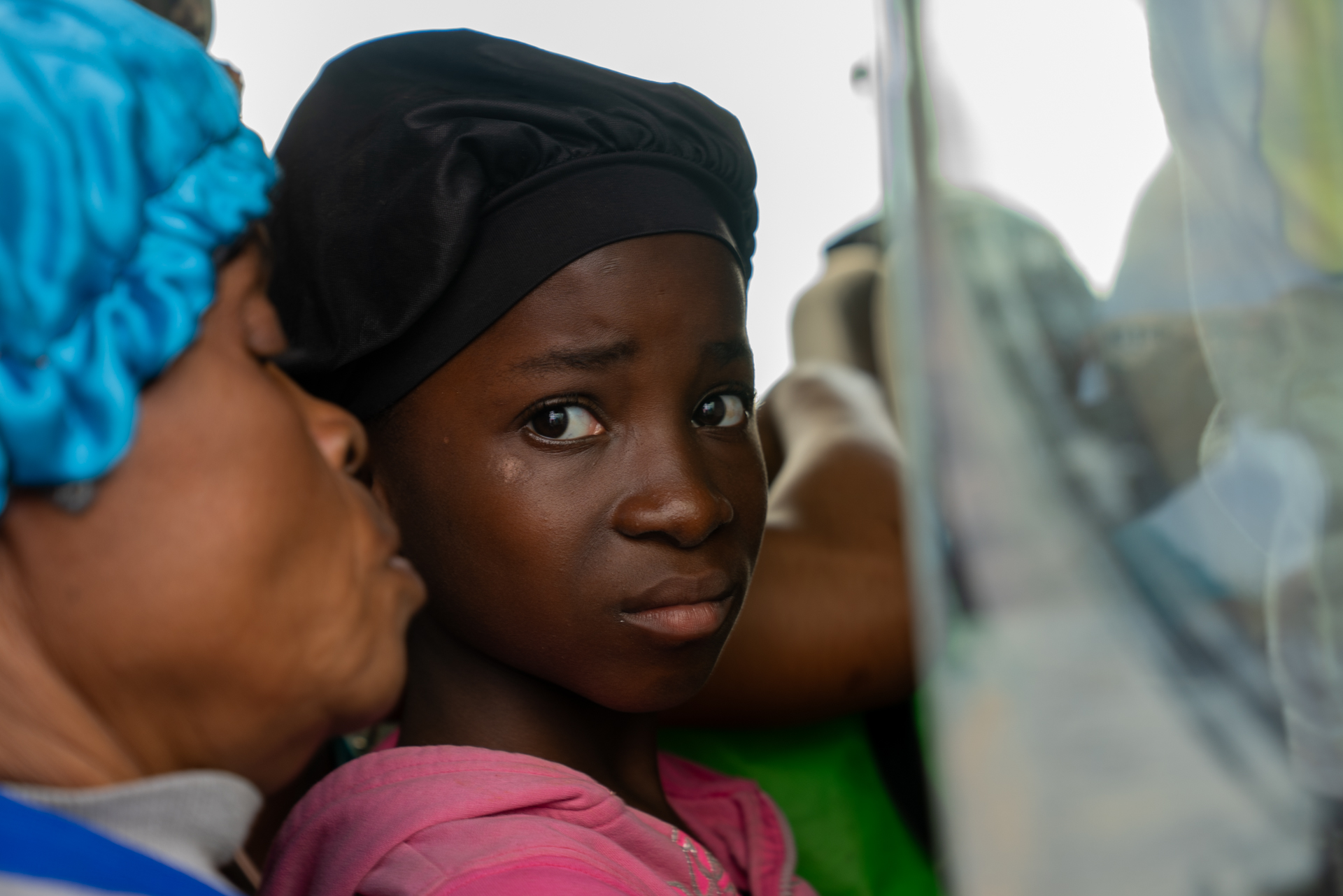 A close-up of a young woman's face as she looks sideways with a tense, uncertain expression, pressed close to another person partially visible at the left edge of the frame. The image is a close-up of a van transporting Haitian deportees to the border.