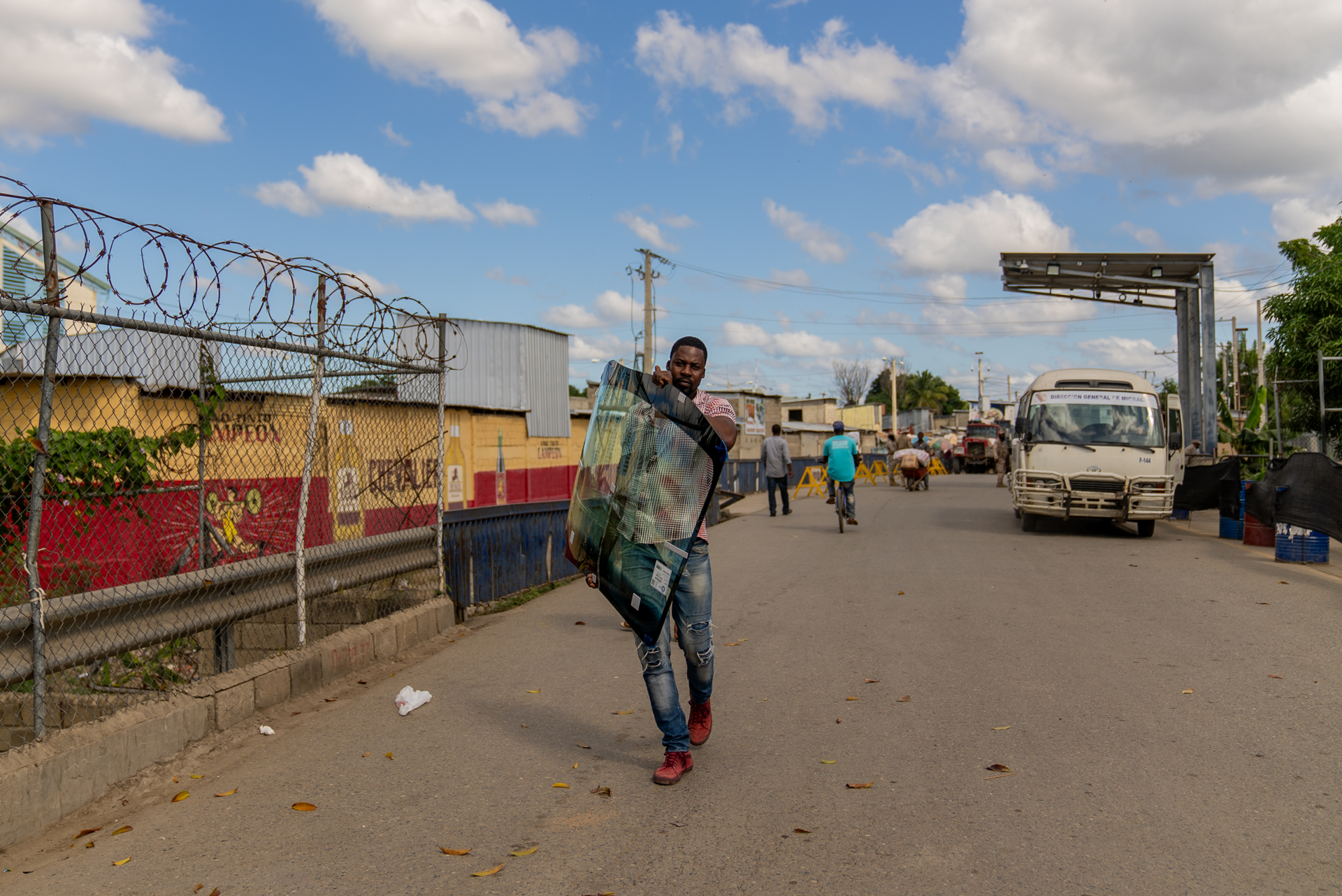 A man carries a large flat-screen TV on his back along a road at the Haiti-Dominican Republic border crossing, glancing back at the camera. Razor wire tops a chain-link fence on the left; a customs gate and a bus are visible in the background.
