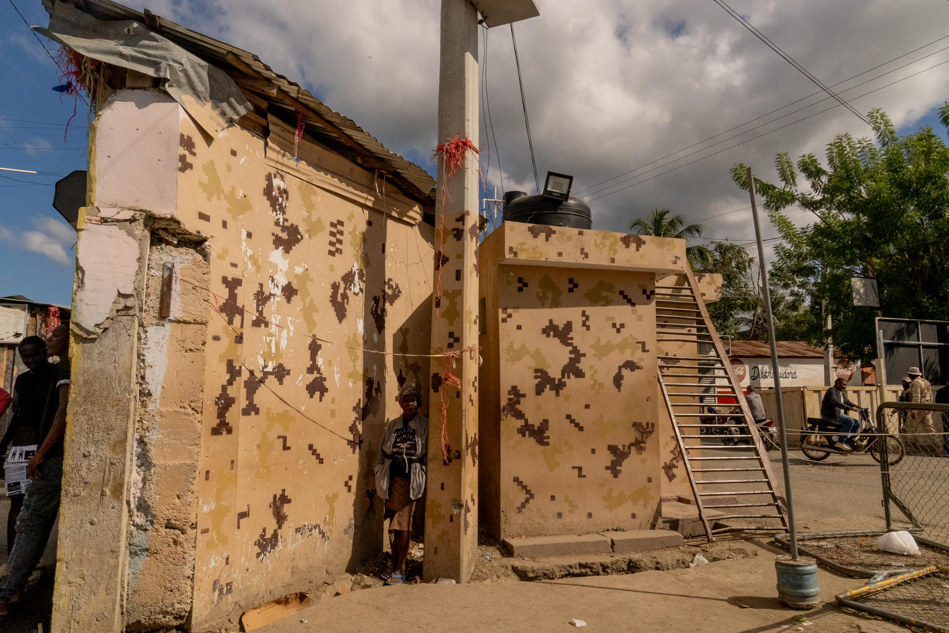 A persone stand in a narrow gap between buildings painted in a camouflage pattern at a border checkpoint. A surveillance light and water tank are mounted on a concrete post; motorcycles and pedestrians pass in the background.