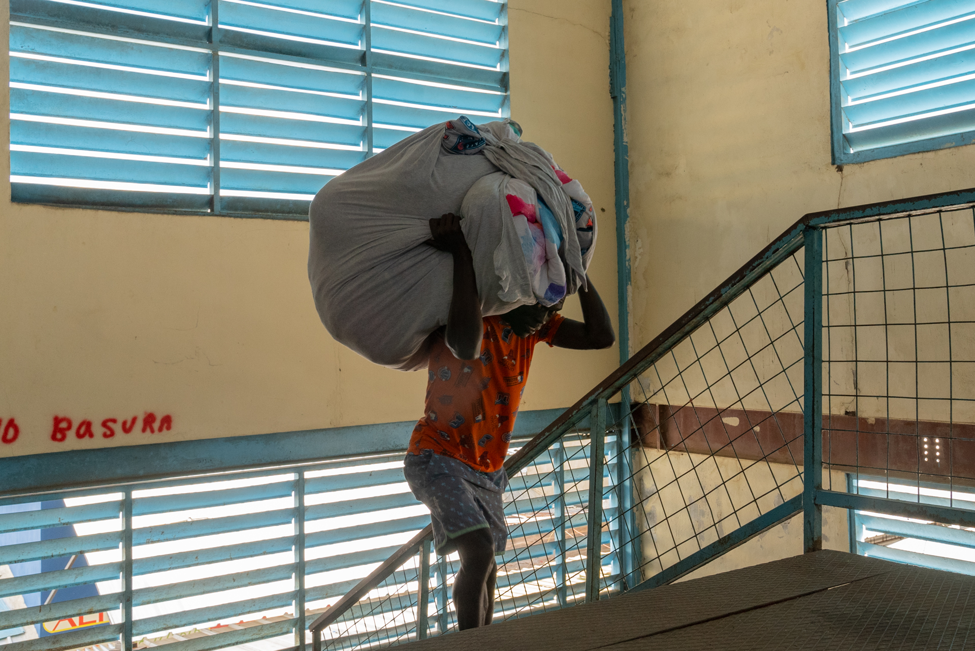 A person in an orange shirt carries an enormous bundle of clothing or textiles on their head and shoulders up a staircase inside a building, nearly obscured by the load. The words "No Basura" are painted in red on the wall beside them.