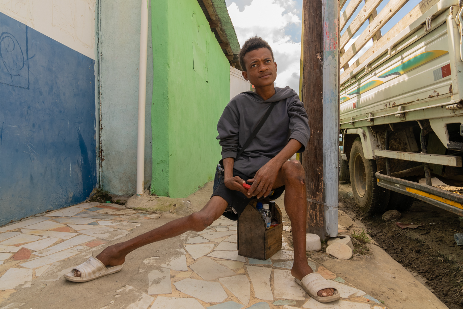 A young man sits on a small wooden shoe-shine box on a tiled sidewalk, one leg extended, looking upward with a watchful expression. Colorful painted walls flank him on the left; a truck is parked to his right.