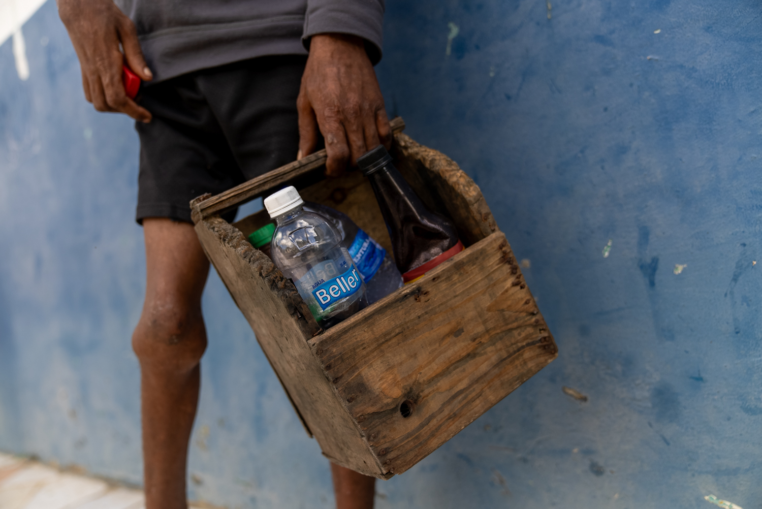A close-up of a worn wooden shoe-shine kit being held at the side, containing a plastic water bottle labeled Beller and a dark glass bottle. The holder's hands are visibly weathered.