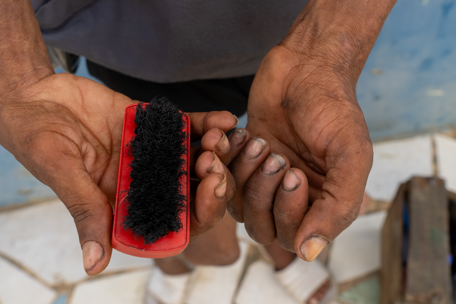 A close-up of two weathered hands holding a small red shoe-shine brush with black bristles. The fingernails are darkened with polish; the hands are cupped open as if presenting the tool.