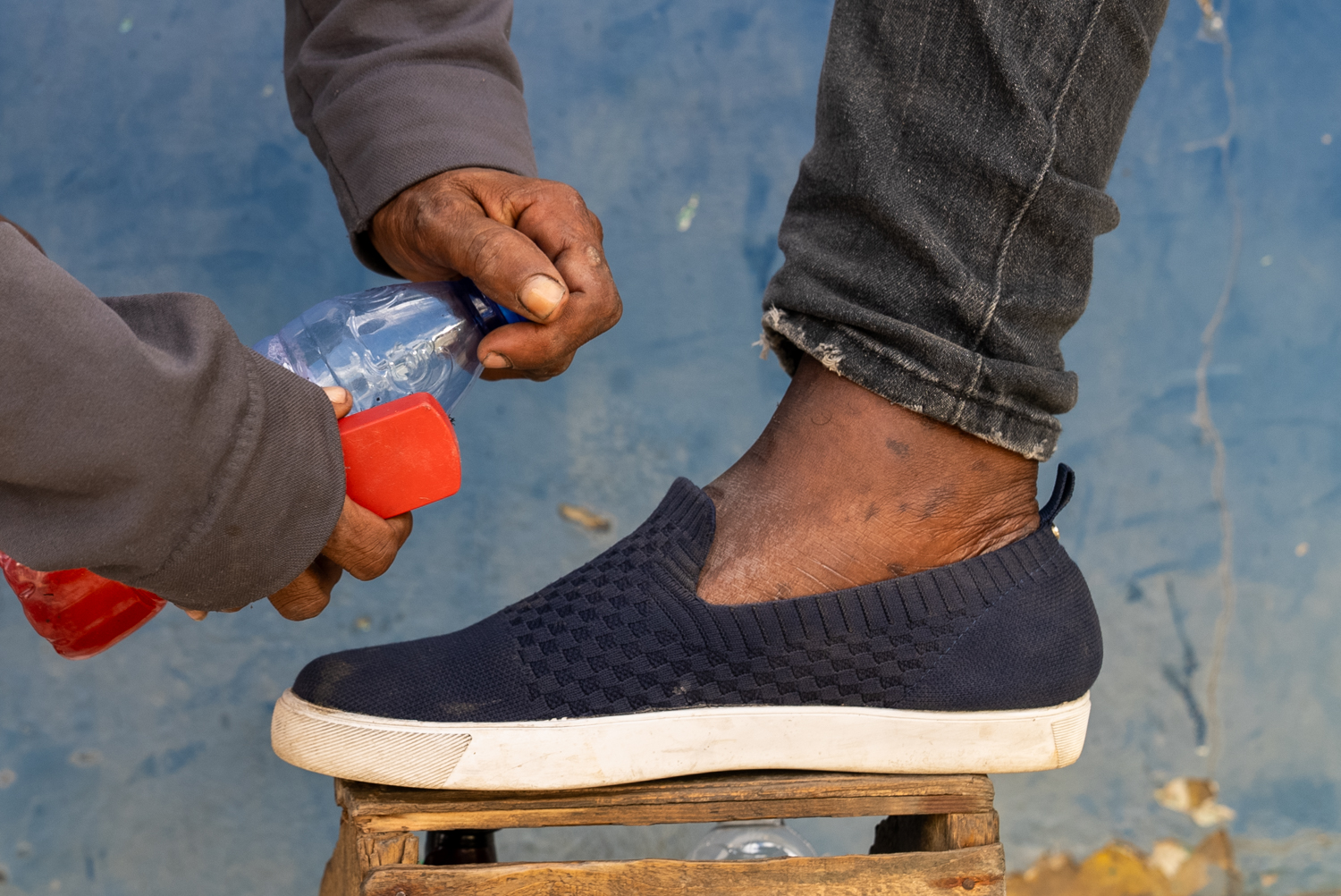 A shoe shiner applies liquid from a small red bottle to the toe of a navy slip-on sneaker resting on a wooden foot rest. The shiner's sleeve is pulled back; a blue painted wall is visible in the background.