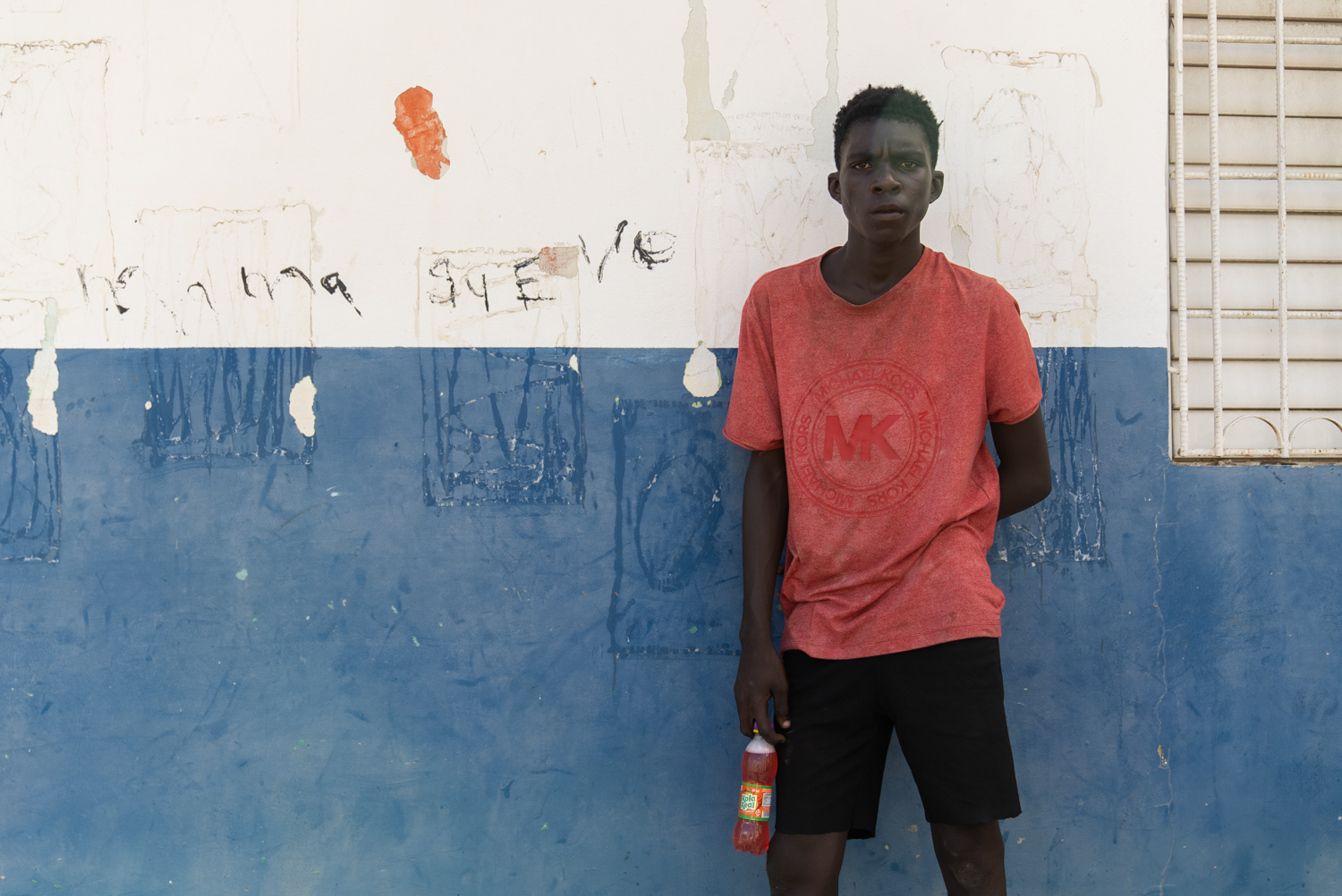 A teenage boy in a red Michael Kors t-shirt stands against a two-tone blue and white painted wall, holding a soda bottle at his side, looking directly at the camera with a guarded expression. Handwritten graffiti marks the wall behind him.