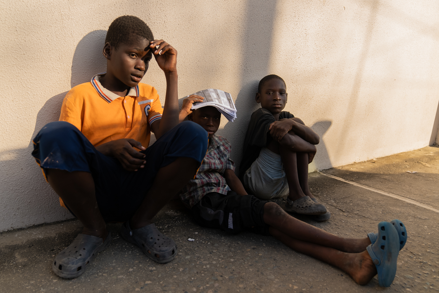 Three boys sit against a sunlit concrete wall in the late afternoon, wearing Crocs and casual clothes. The boy on the left in an orange polo looks toward the camera; the middle child shields his face with a folded paper; the boy on the right stares forward with arms wrapped around his knees.
