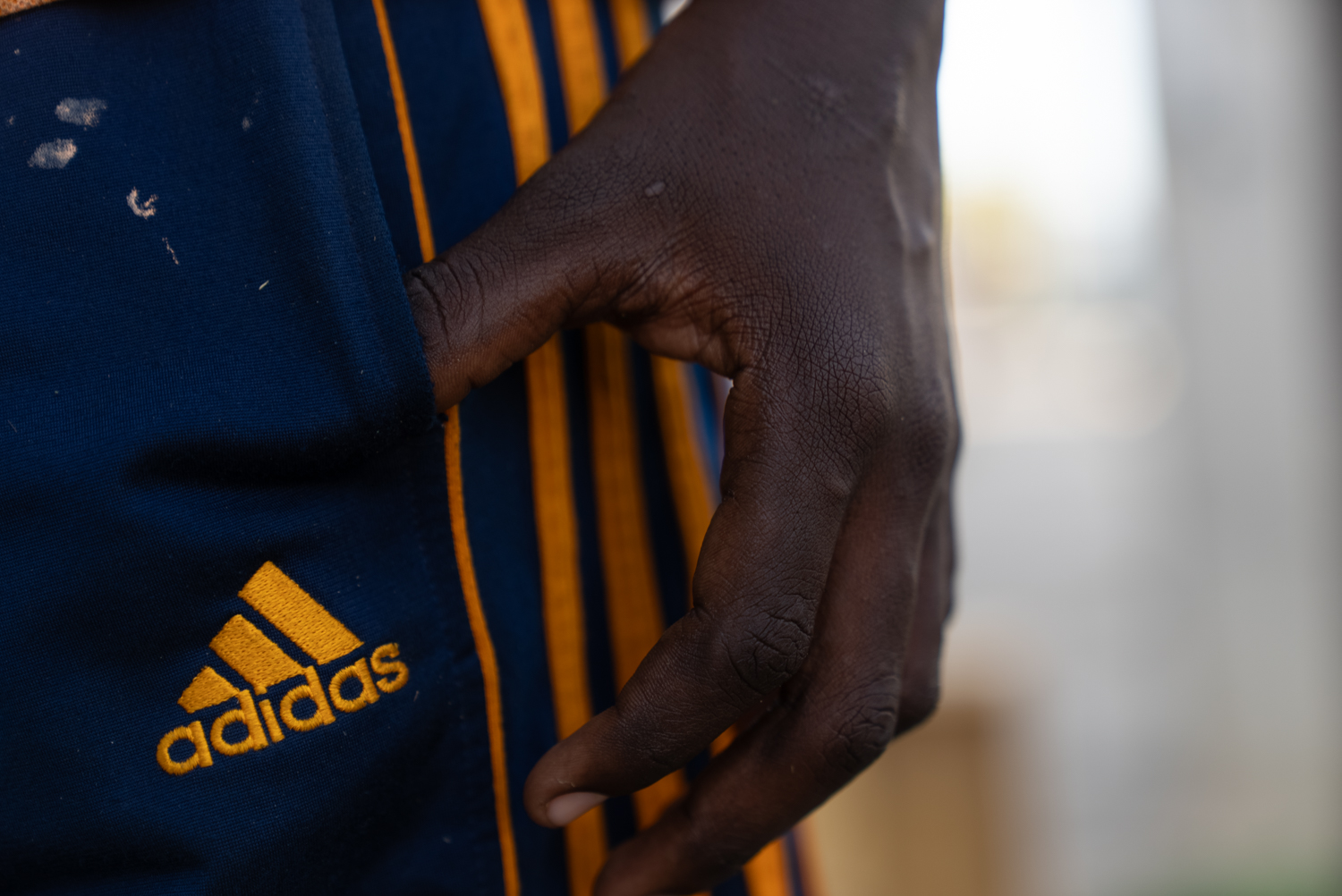 A close-up of a person's hand resting against their hip on navy blue Adidas track shorts with gold stripes. The hand is calloused and worn; paint or polish stains mark the fabric.