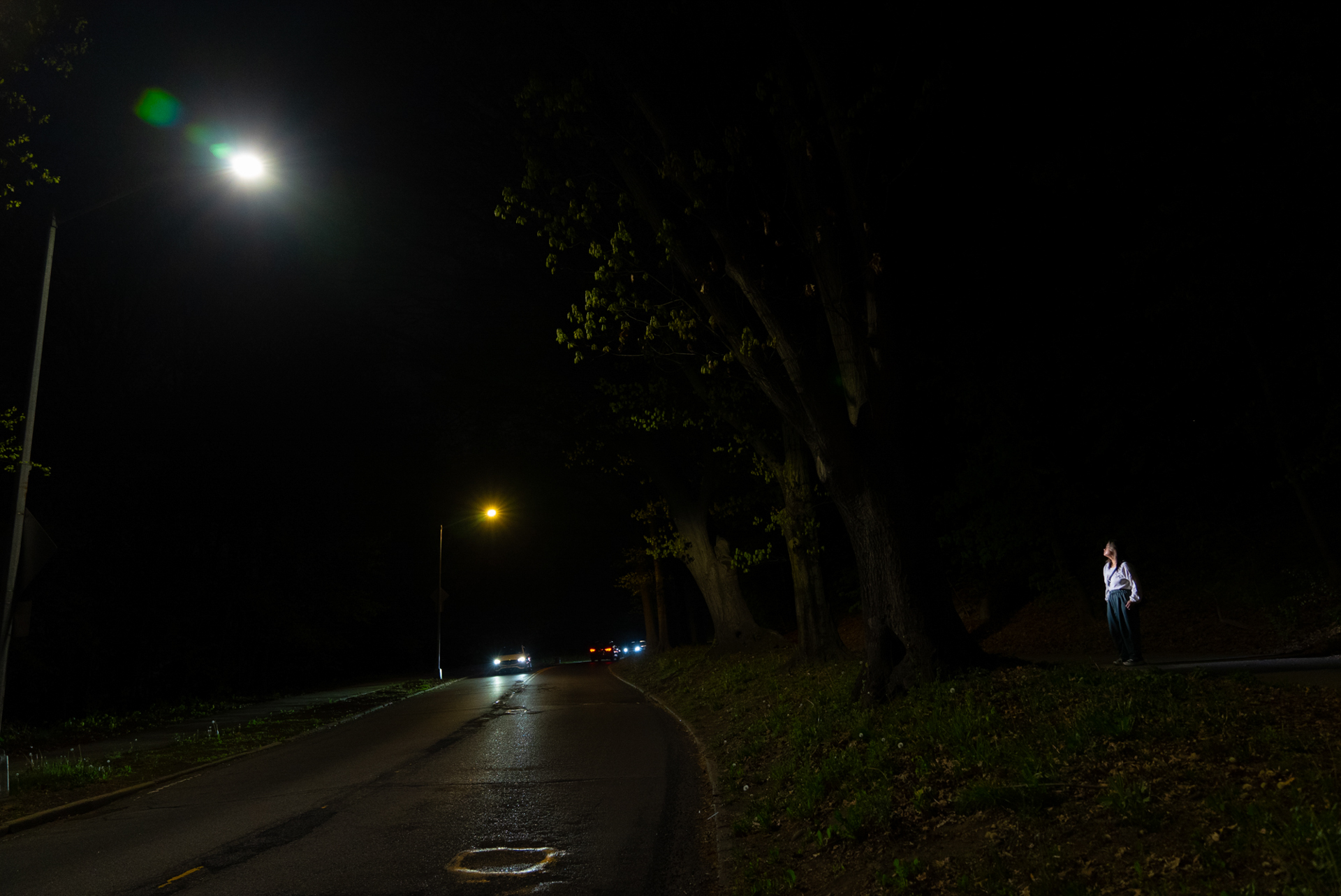 A woman in a white shirt stands alone on the shoulder of a dark road beside a tree-filled park at night, lit by a single distant streetlight. Car headlights approach in the distance; large trees line the right side of the road