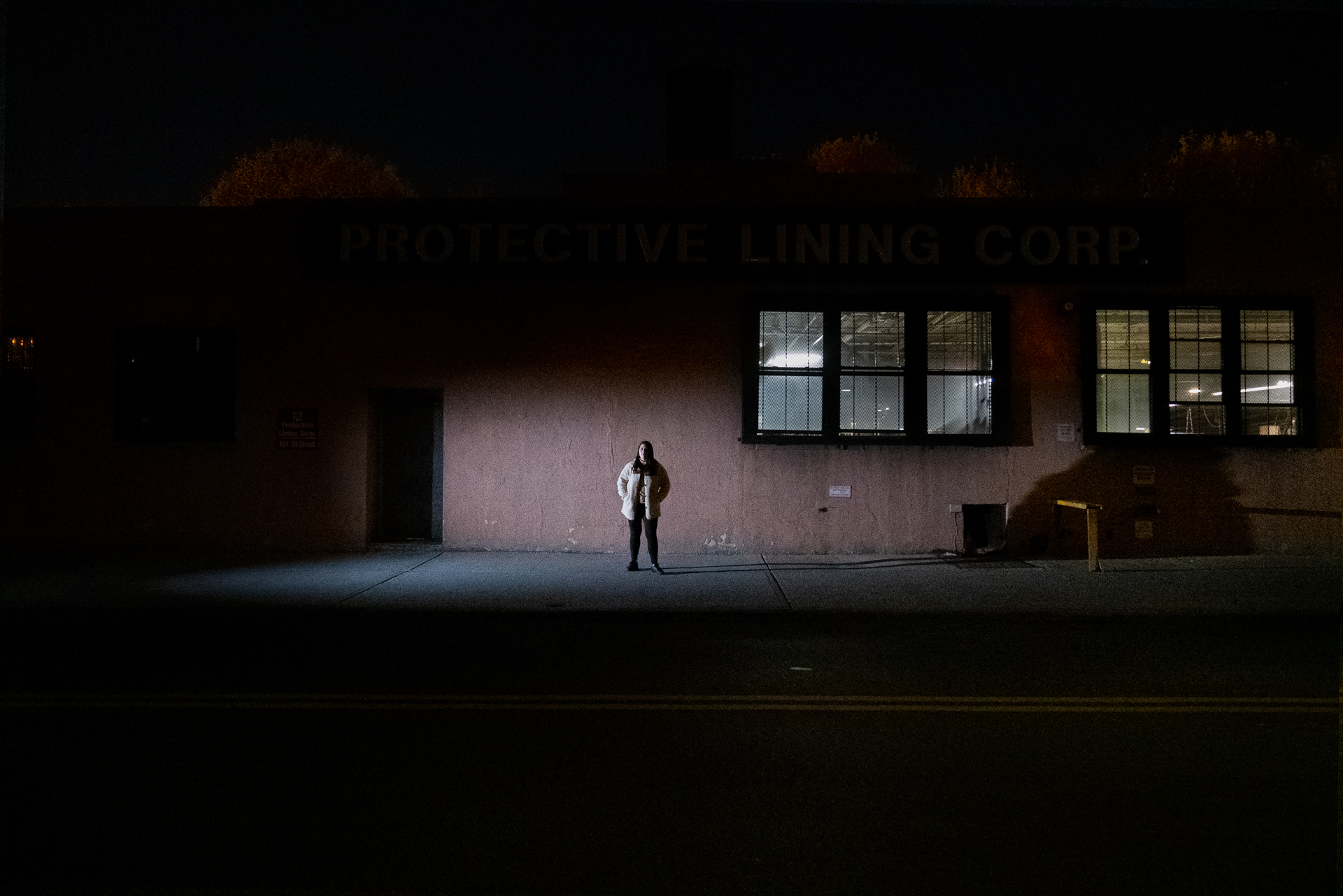 A woman in a white coat stands centered against the stucco façade of an industrial building at night. Interior lights glow through factory windows behind them; the surrounding sidewalk is in near-total darkness.