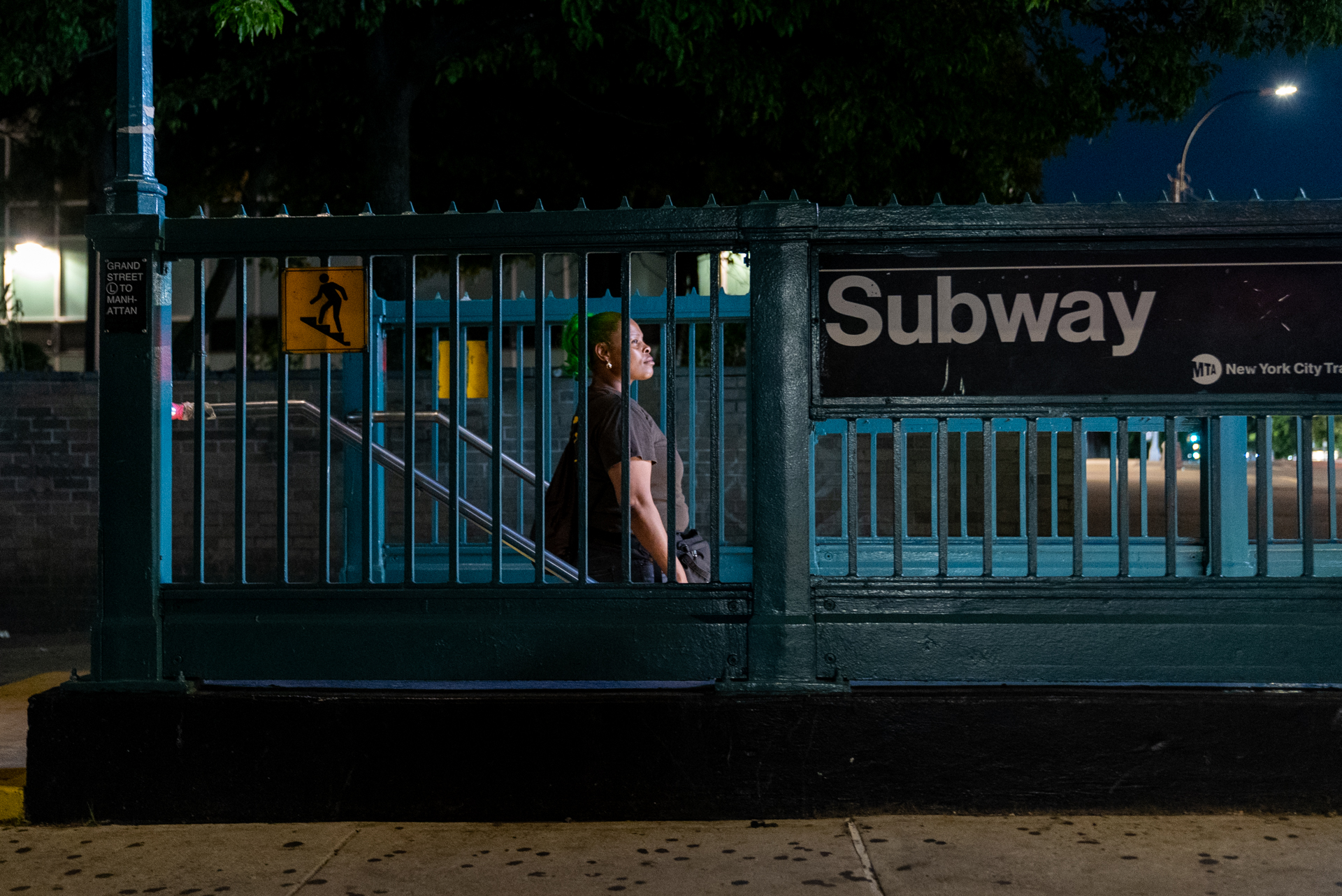 A woman in a UPS uniform of a brown T-shirt and shorts pauses mid-stride on the staircase of a New York City subway entrance at night, viewed through the green iron fence. An MTA sign reads "Subway / Grand Street to Manhattan
