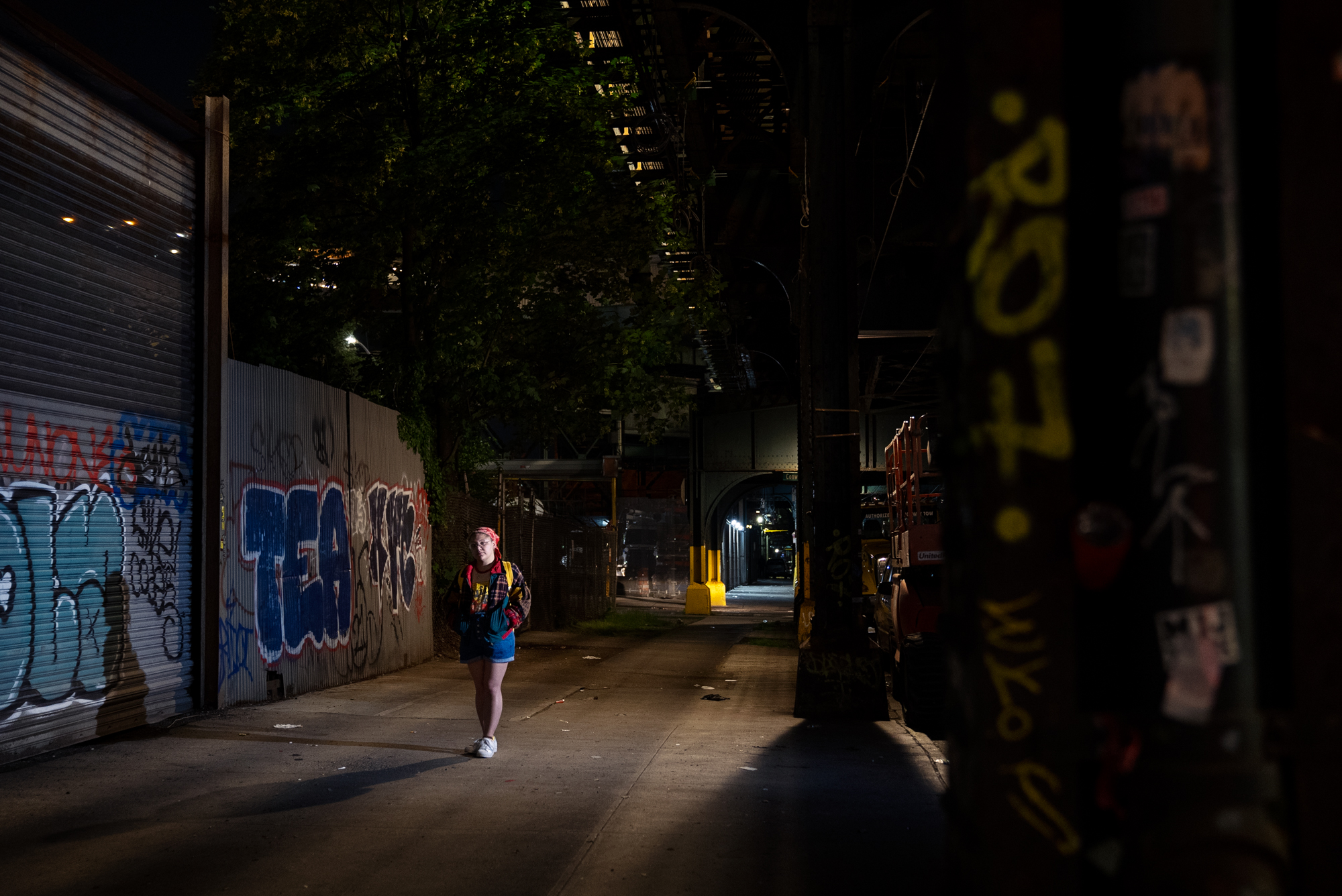 A young woman wearing a red bandana over her hair and a backpack walks alone at night through a narrow passage beneath an elevated structure, flanked by corrugated metal shutters and graffiti-covered walls. City lights are visible beyond an archway in the distance.