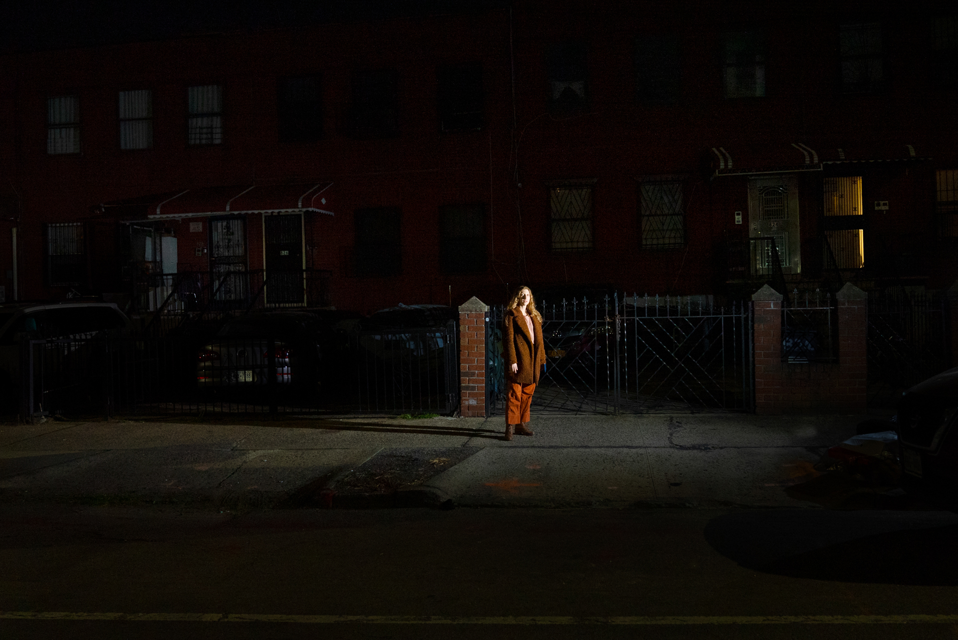 A woman in a long brown coat and rust-colored trousers stands alone on a sidewalk at night, lit by a single off-camera light. Dark red-brick apartment buildings and iron gates line the block behind her.