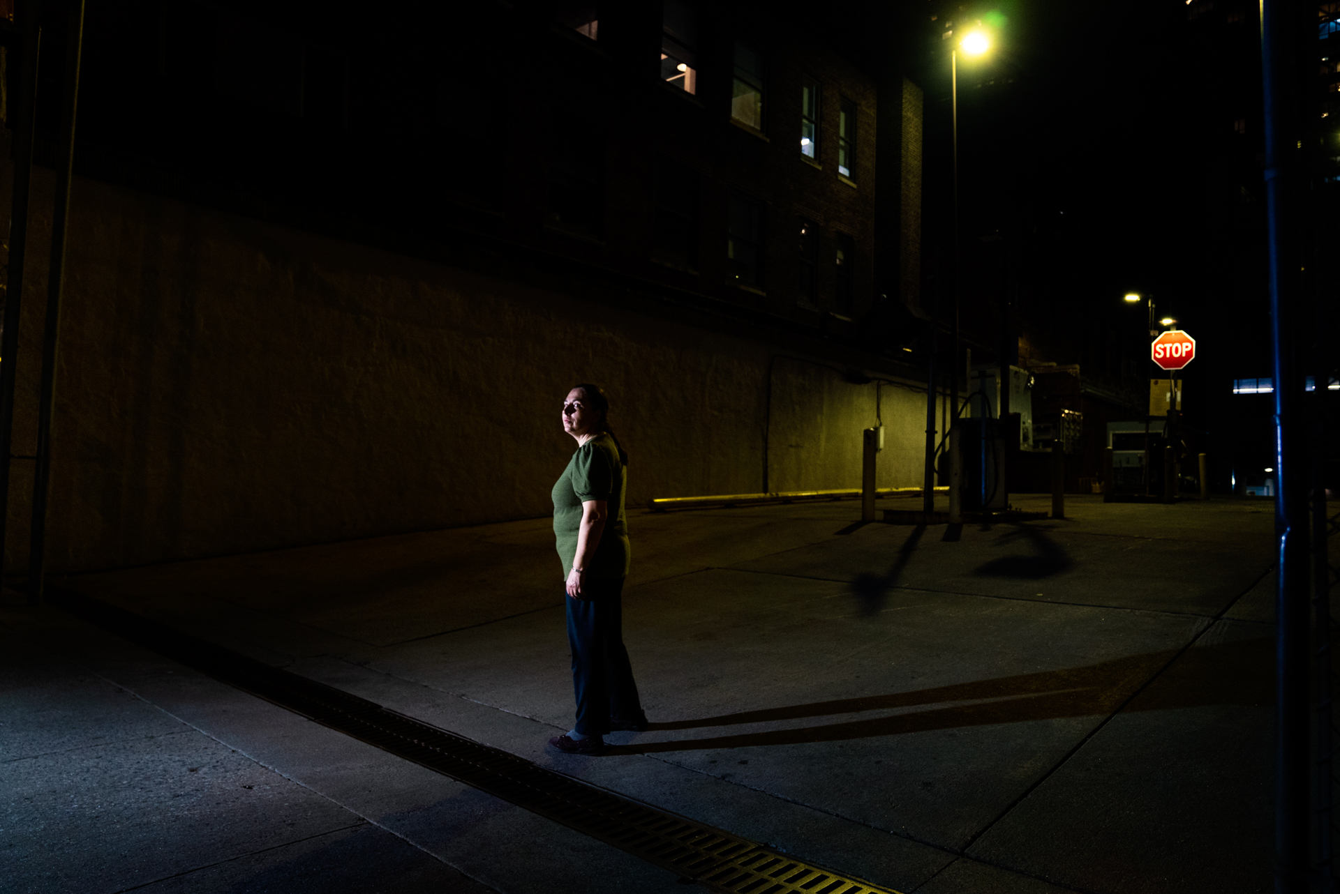 A woman in a green shirt and glasses stands on a sidewalk at night beside a large blank industrial wall, lit from above. A stop sign glows red in the background; yellow light warms the loading dock area behind them.