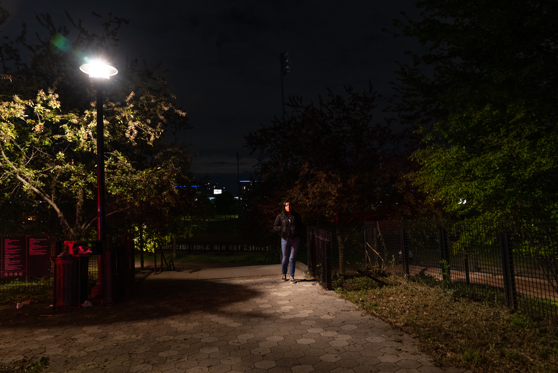 A young woman in a dark jacket stands on a paved park path at night, between an illuminated park lamp and a row of iron fencing. Trees frame the scene; city lights are faintly visible on the horizon.
