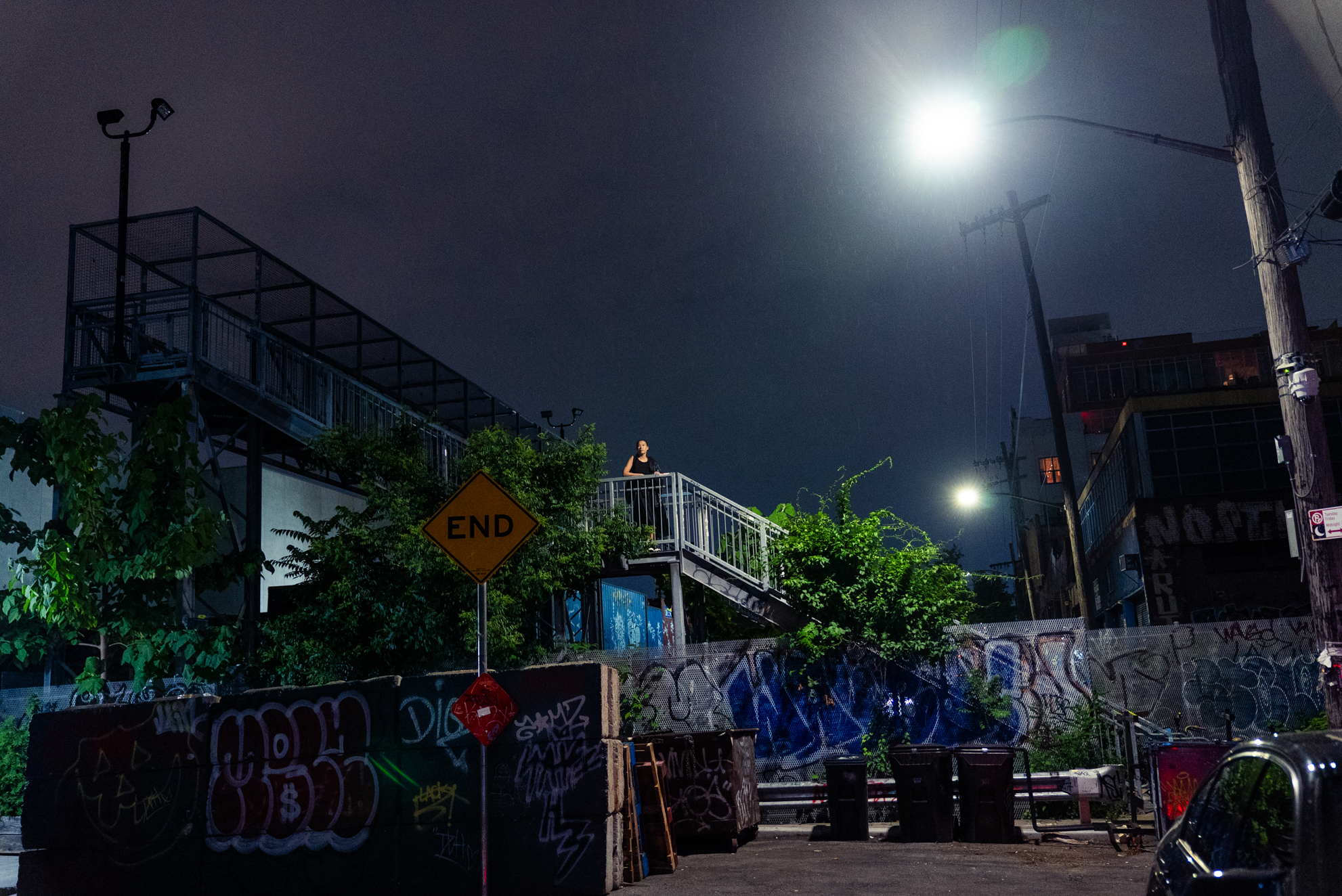 A woman stands on an elevated metal staircase landing at night in an urban alley, lit from below against a dark sky. Graffiti-covered walls and a yellow diamond "END" road sign frame the scene; streetlights glow in the background.