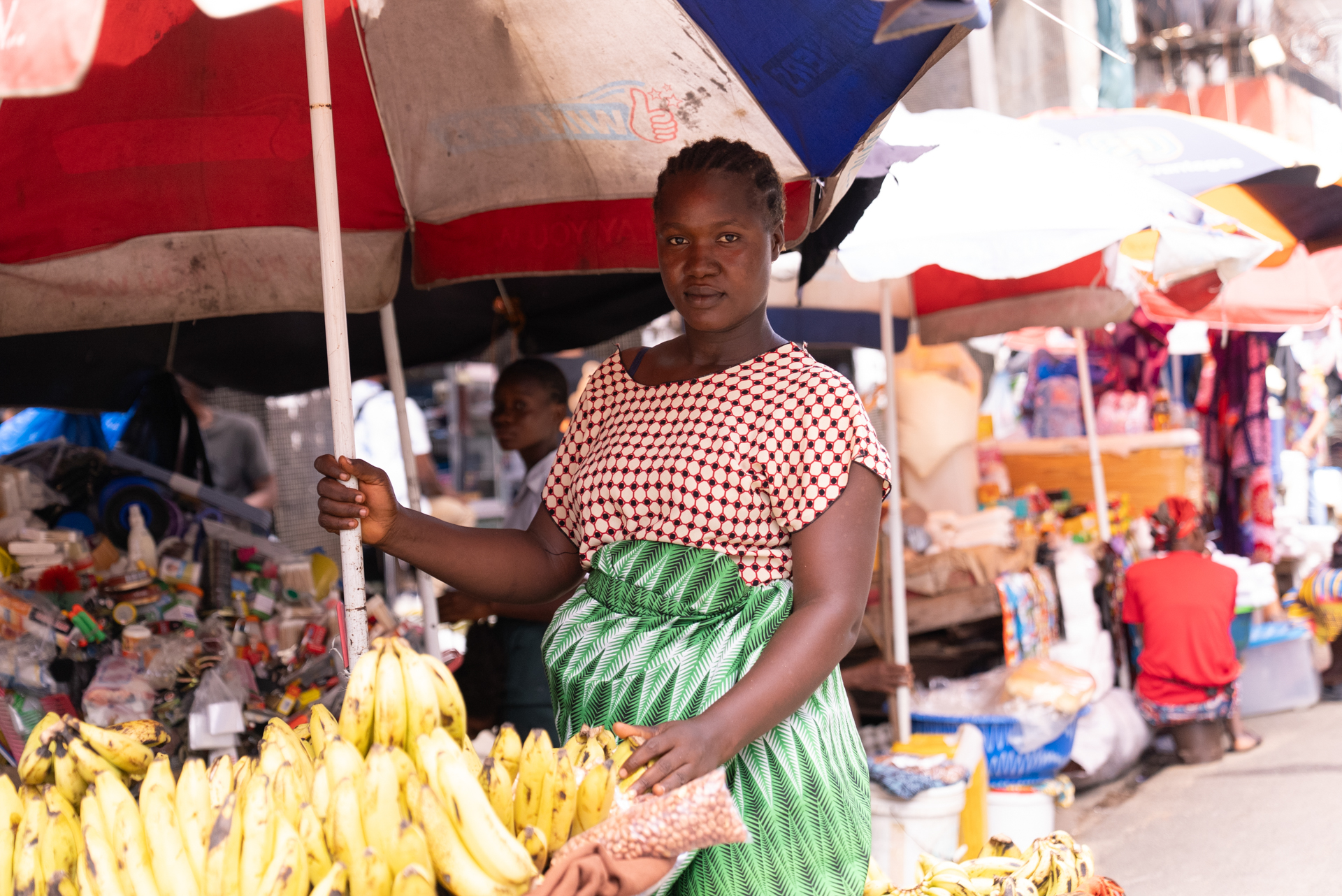 A pregnant woman in a patterned red-and-white top and green leaf-print skirt stands at a banana stall in a busy outdoor market, holding a parasol pole and looking directly at the camera.