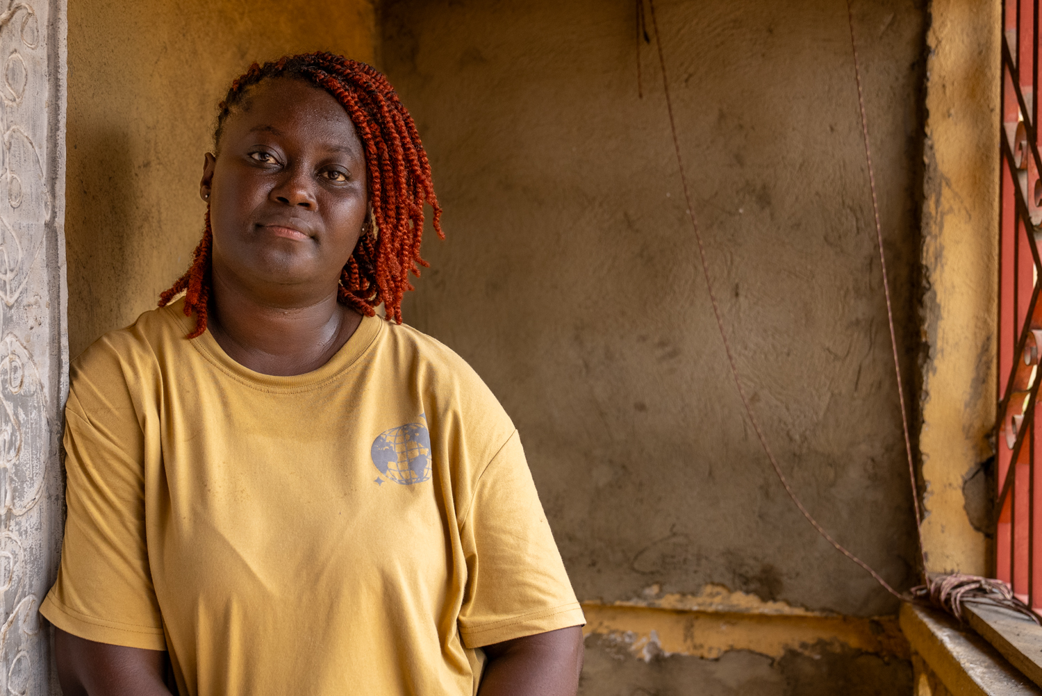 A woman with long red braids and a yellow t-shirt leans against a concrete wall, looking slightly upward with a calm expression. Warm daylight catches her face; a painted column and a red-barred window are visible beside her.