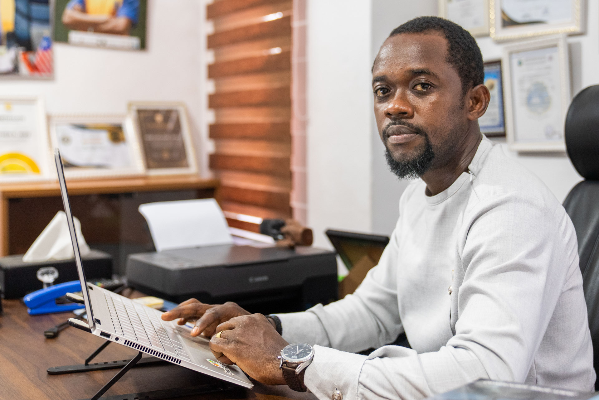 A man in a white collarless shirt sits at a desk with a laptop, looking toward the camera, surrounded by framed certificates and awards on the wall behind him.