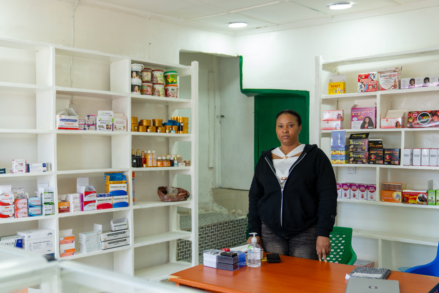 A woman stands behind a counter in a small pharmacy, surrounded by white shelves; about a third are bare and two thirds are stocked with medications, supplements, and health products. She faces the camera directly with a neutral expression.