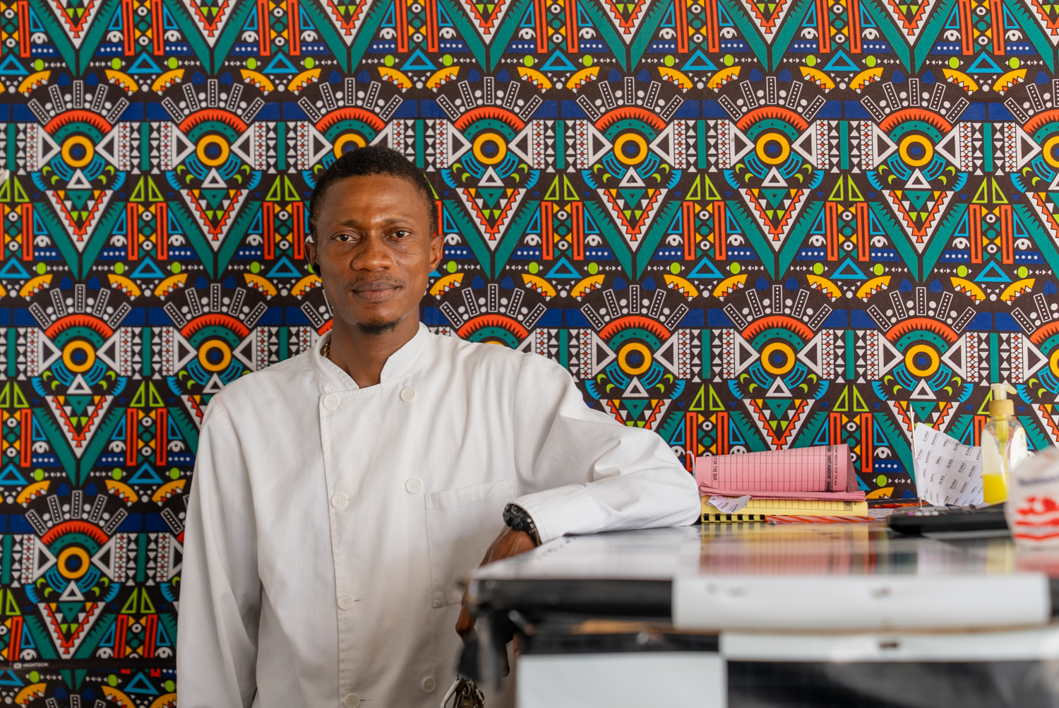 A man in a white chef's coat leans against a counter, looking directly at the camera. Behind him, a wall is covered in bold, colorful African wax-print fabric in geometric patterns of teal, orange, yellow, and red.