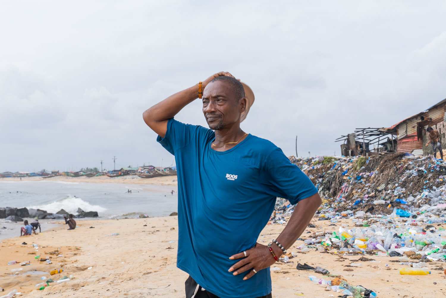 A man in a blue t-shirt stands on a littered beach, one hand on his hip and the other raised to hold his hat against the wind. Behind him, plastic waste and debris pile up along the shoreline beside weathered wooden structures; ocean waves and a fishing community are visible in the distance.