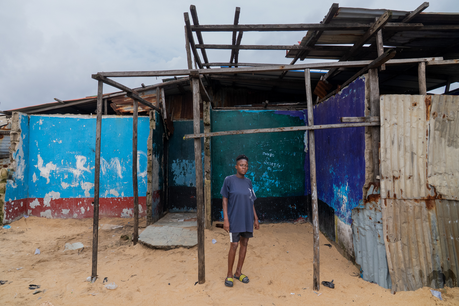A woman stands beneath the skeletal wooden frame of a storm-damaged structure on a sandy beach. The walls behind her are painted blue, green, and purple; corrugated metal panels line the sides.