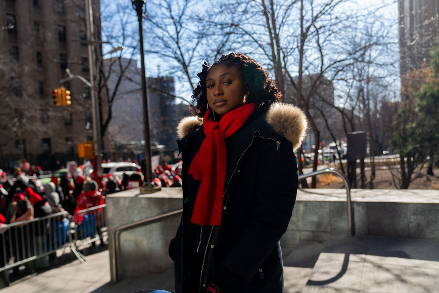 A woman in a red scarf and fur-trimmed coat stands at a winter rally in New York City, protesters in red visible behind her