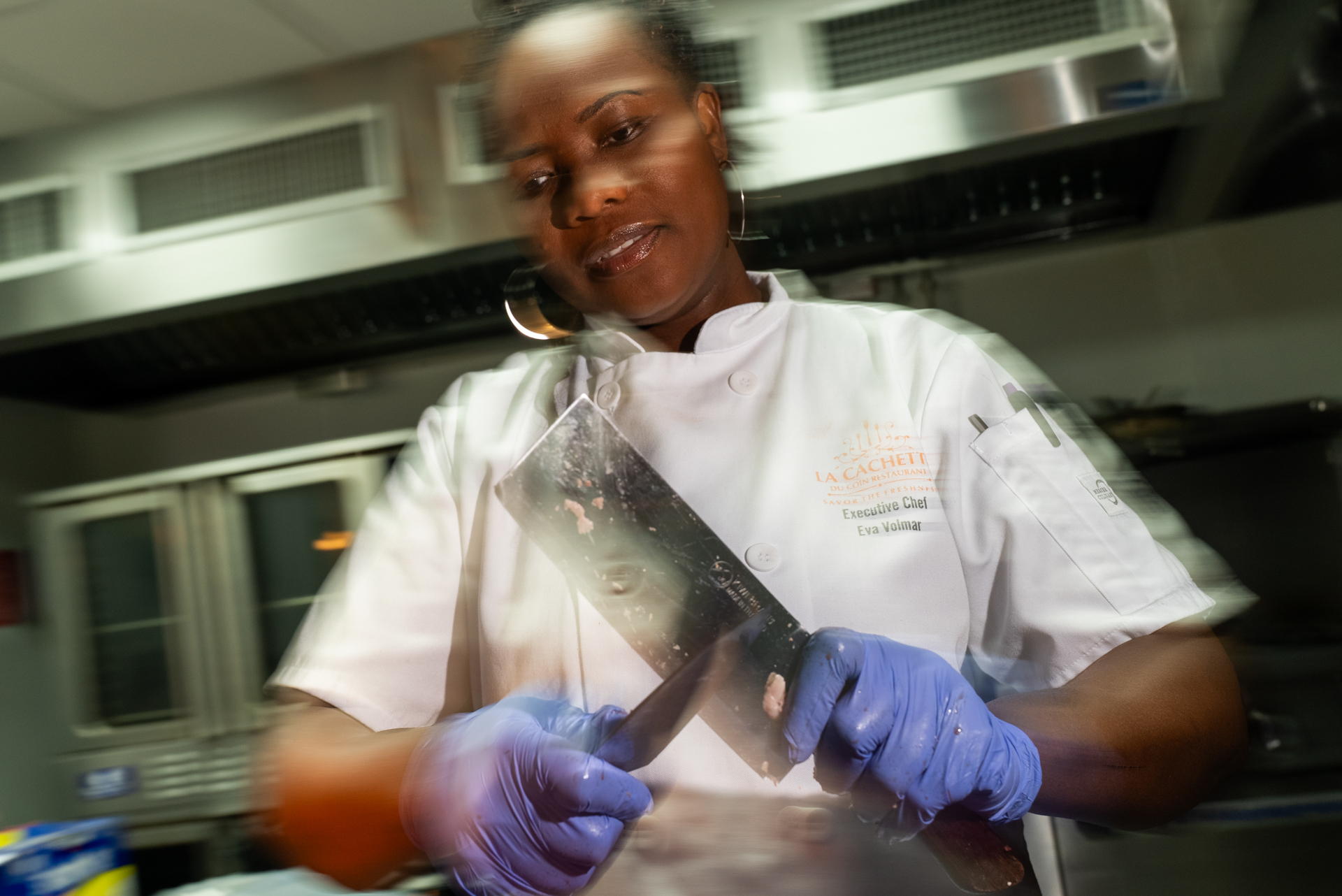 Executive Chef Eva Volmar holds a cleaver in blue latex gloves in a professional kitchen, her white chef's coat bearing the La Cachette restaurant logo, the image shot with a motion-blur effect from a low angle