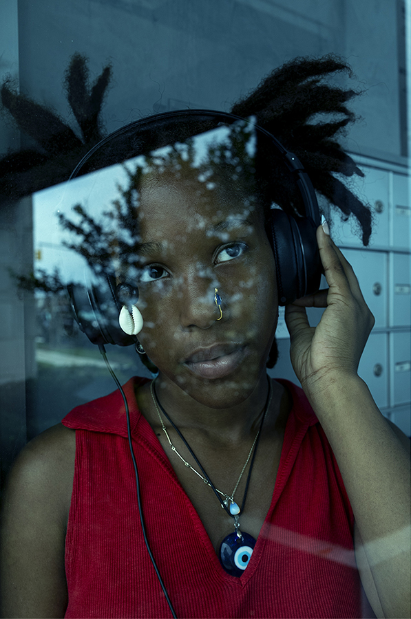 A woman in a red sleeveless top holds headphones against her head, a tree reflection visible through the glass behind her