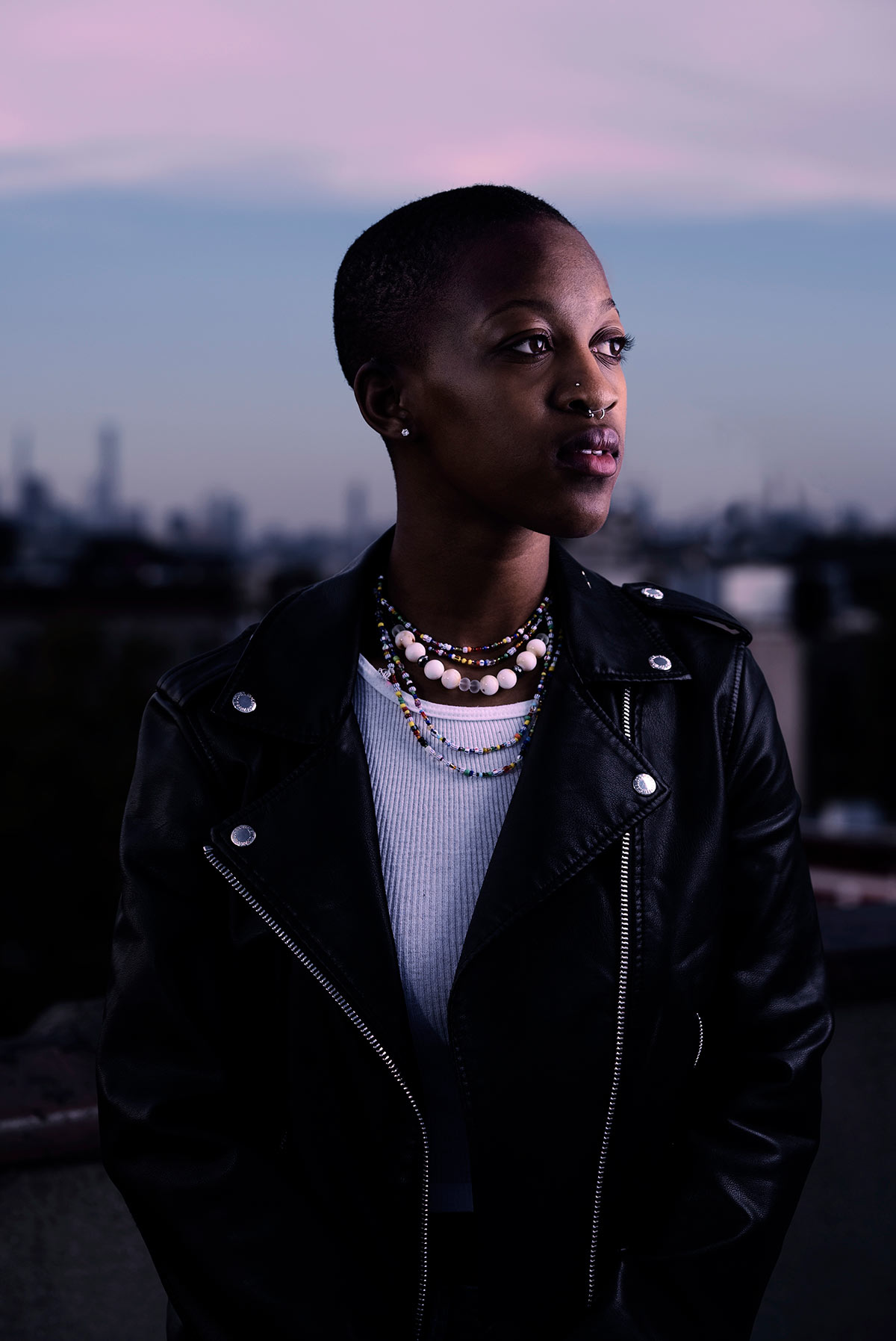 Portrait of a woman in a leather jacket with layered bead necklaces against a dusky city skyline