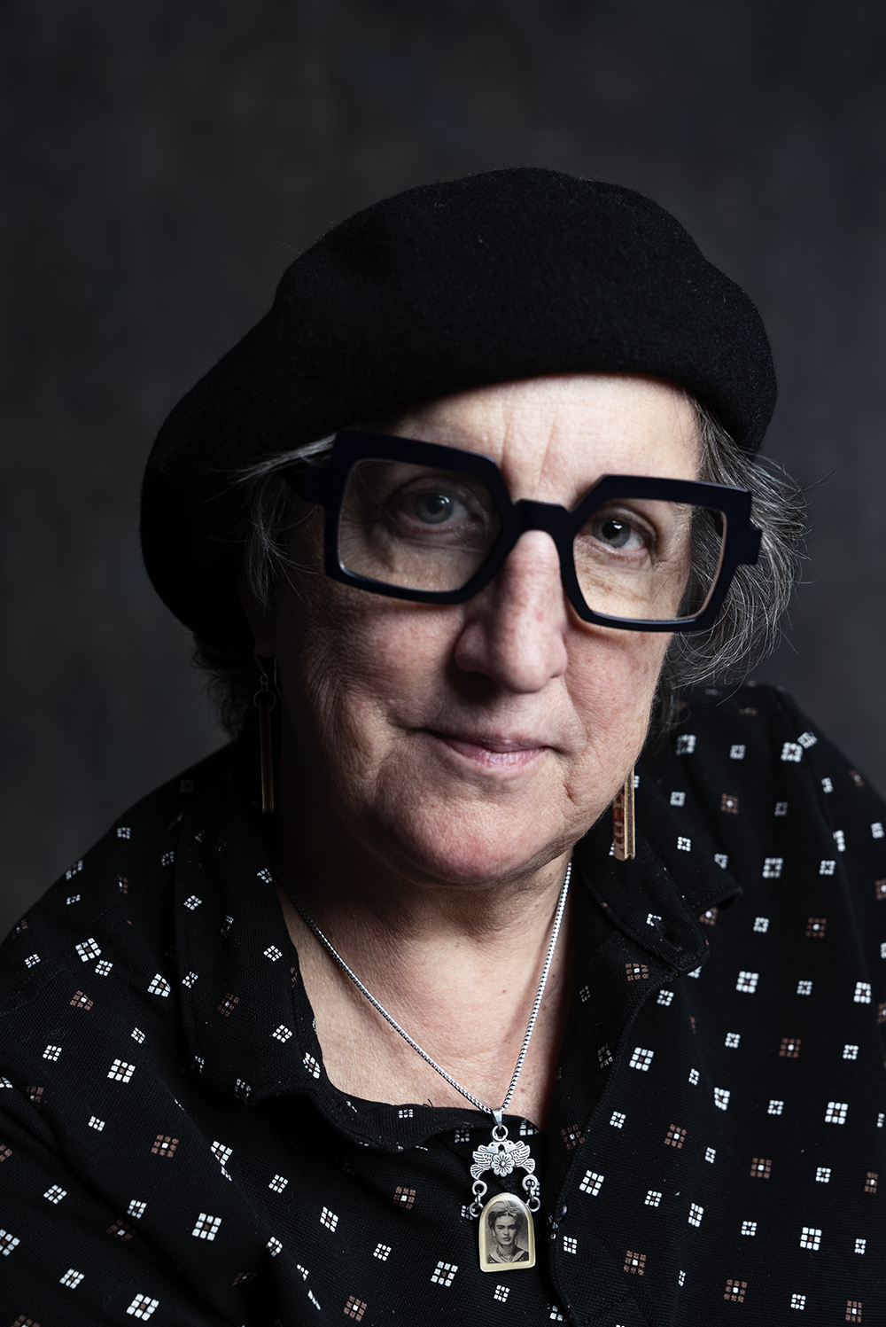 Close portrait of an older woman in a black beret and bold square glasses with a Frida Kahlo pendant necklace