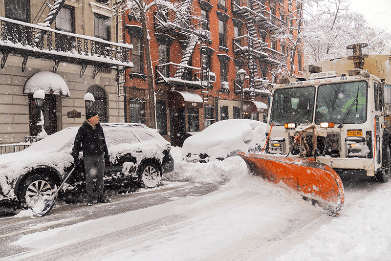 A man shovels snow beside a parked car on a snow-covered Manhattan street.