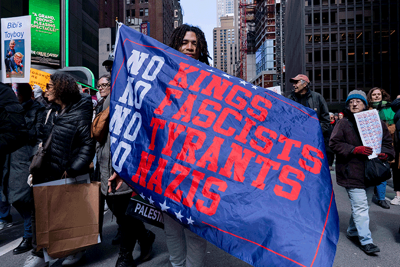 A protester holds a large blue banner reading No Kings Fascists Tyrants Nazis at a midtown Manhattan march.